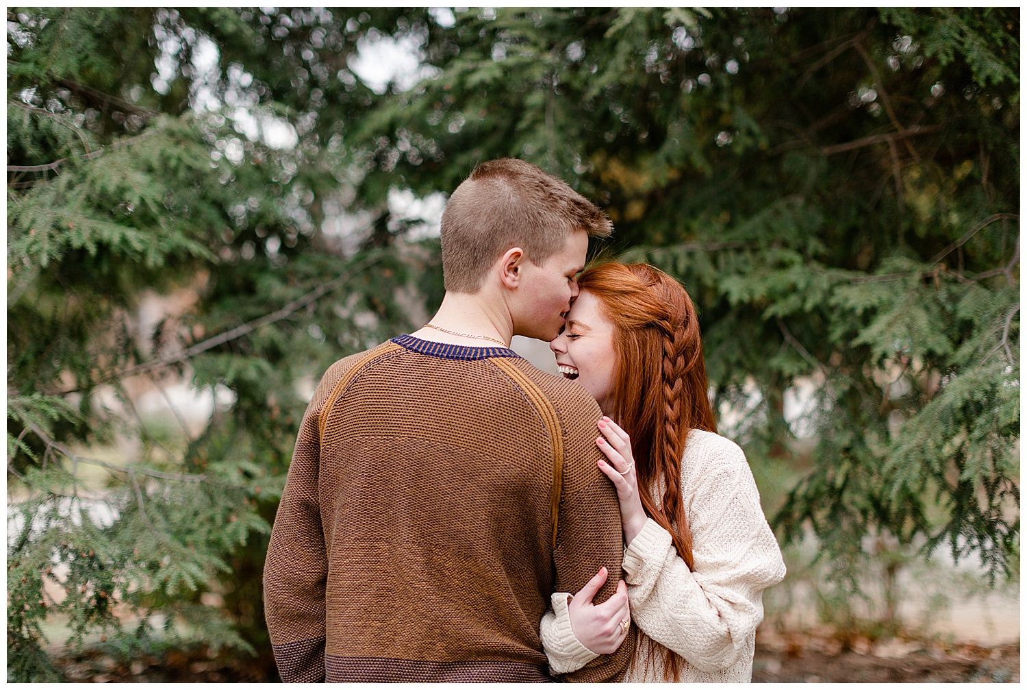 Grant kissing Ashley on the forehead in the evergreen trees during their engagement session at woodward park.